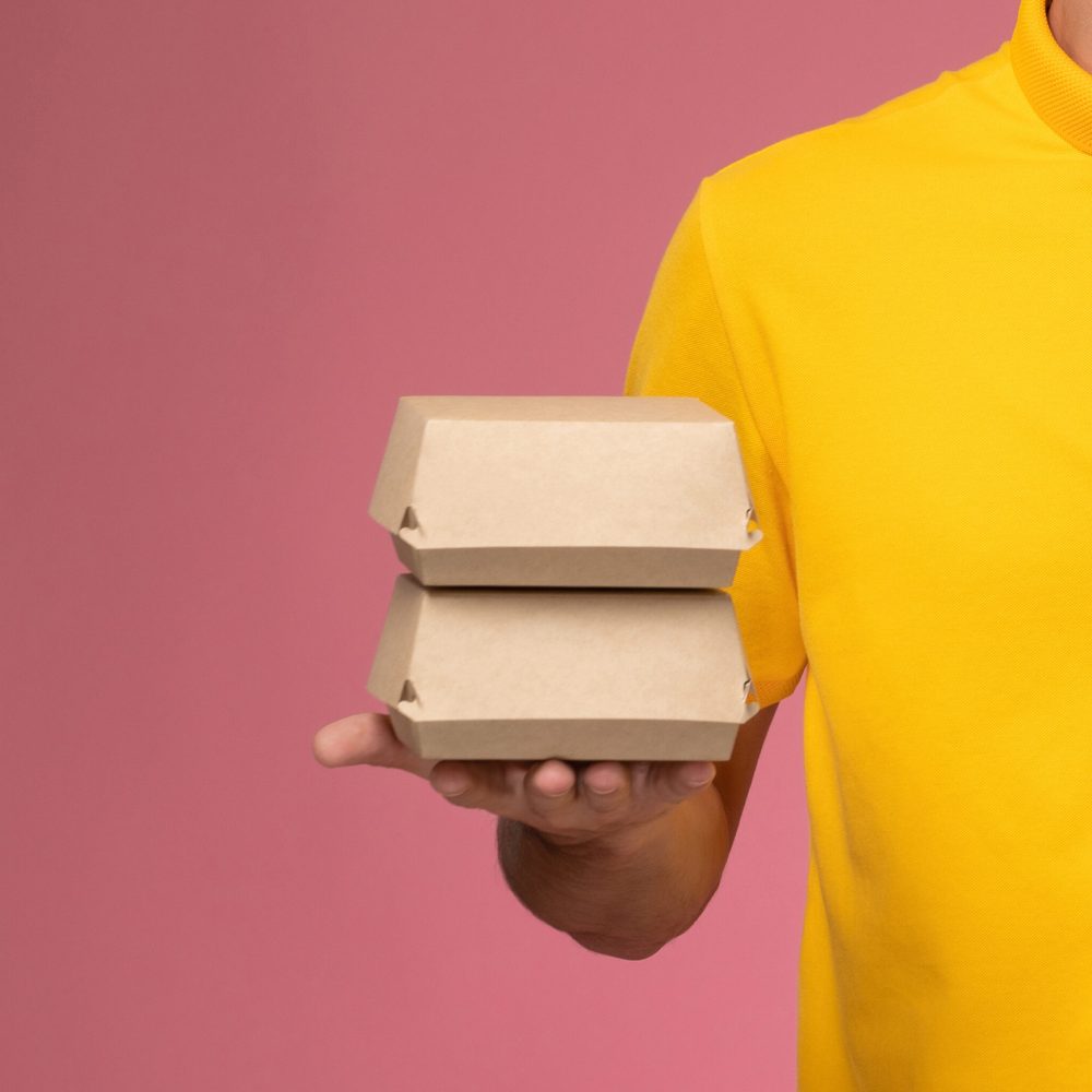 front view male courier in yellow uniform cape with phone and fo front view male courier in yellow uniform cape with phone and food packages on his hands on light-pink background delivering job service uniform